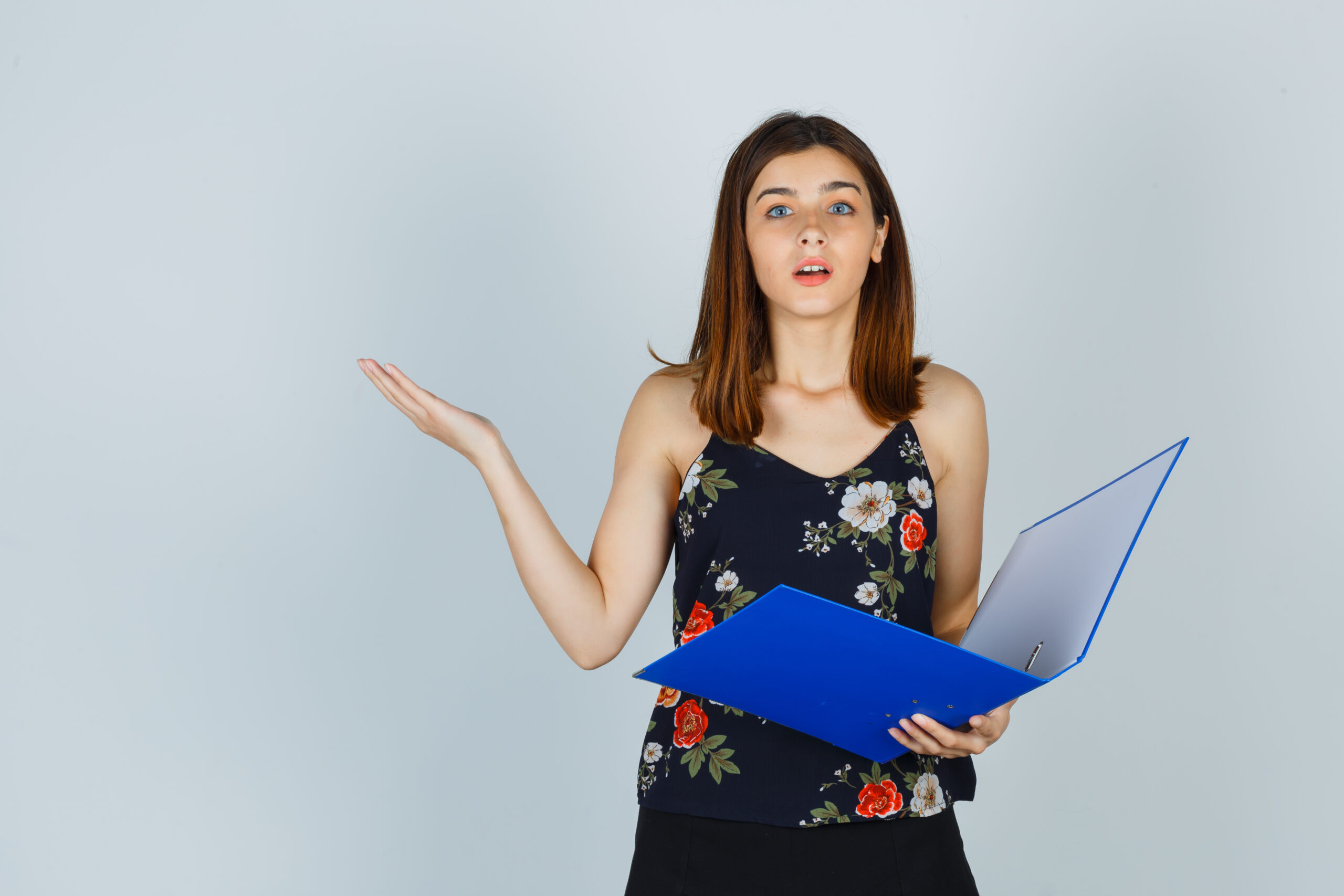 Portrait of young lady holding folder while showing helpless gesture in blouse, skirt and looking puzzled front view
